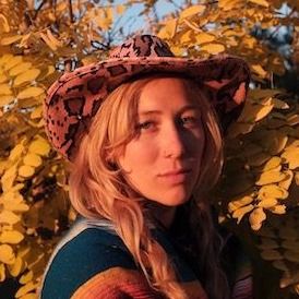 A person with long blond hair, looks at the camera. They are wearing a pink snake skin cowboy hat, and are standing in front of a tree with yellow leaves. The light is warm and glowing.